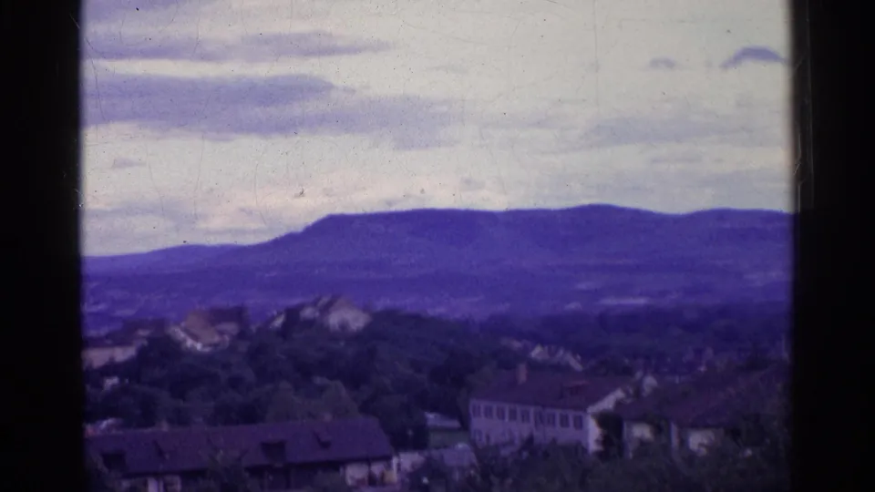 A faded 8mm Swiss landscape frame shows rooftops, trees, and a broad purple mountain ridge beneath a cloudy sky.