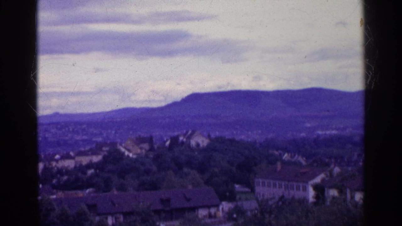 1961 Swiss 8mm frame showing a hillside neighborhood with rooftops, trees, distant mountains, and a cloudy sky.