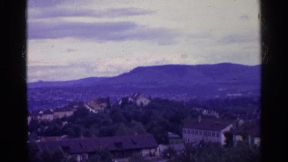 1961 Swiss 8mm frame showing a hillside neighborhood with rooftops, trees, distant mountains, and a cloudy sky.
