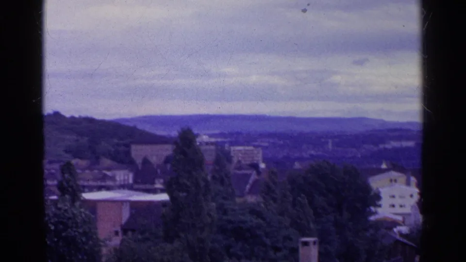 1961 Swiss home-movie frame showing foreground trees, rooftops, low hills, and a distant townscape under overcast light.