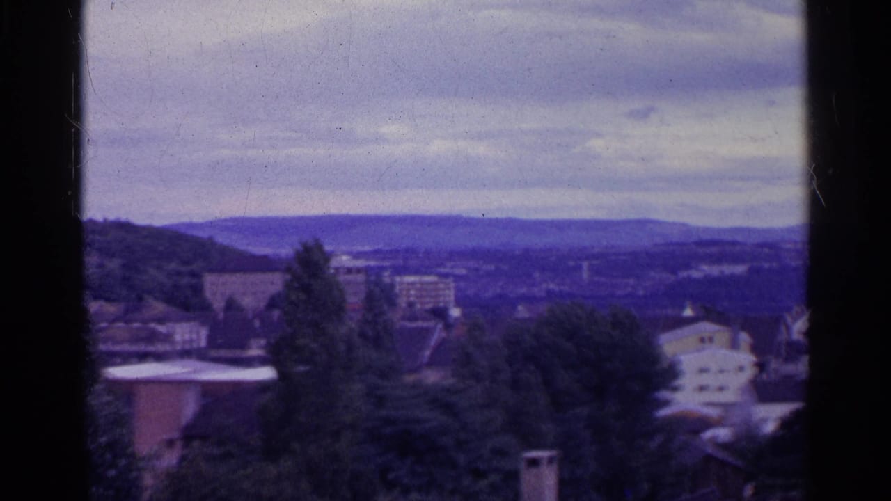 1961 Swiss village view from above, with rooftops, trees, apartment blocks, distant hills, cloudy sky, and dark 8mm frame borders.