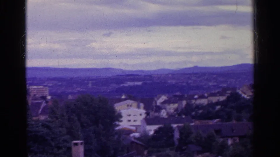 1961 Swiss valley scene with houses, trees, distant low mountains, overcast sky, and soft purple-blue 8mm color shift.