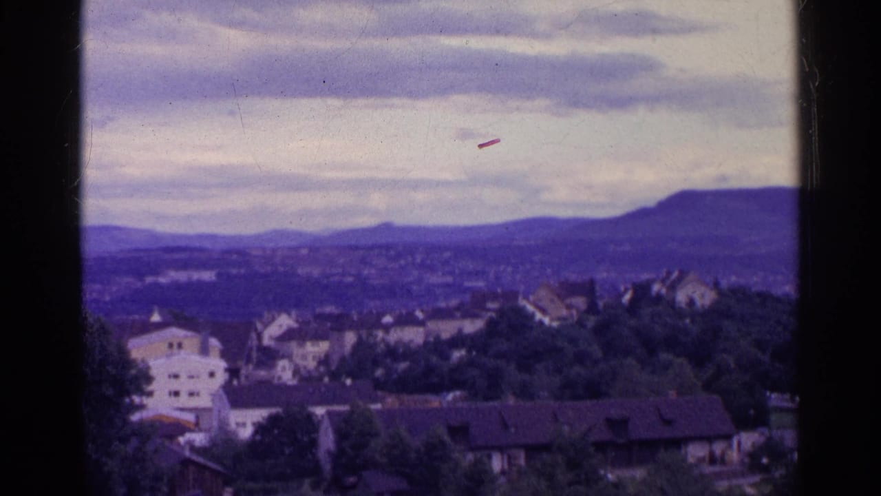 1961 Swiss 8mm frame showing a hillside town, trees, distant ridge, and cloudy sky with a purple-blue color cast.