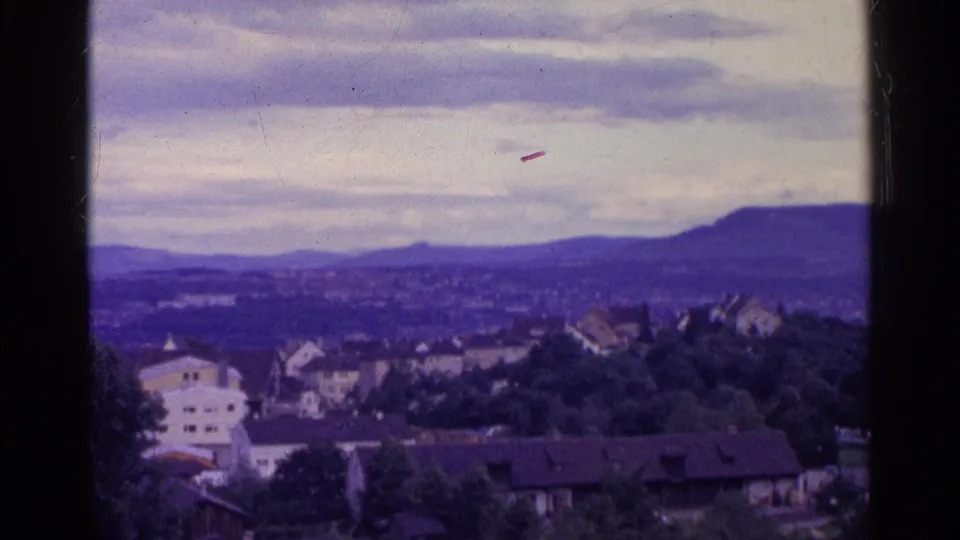 1961 Swiss 8mm frame showing a hillside town, trees, distant ridge, and cloudy sky with a purple-blue color cast.