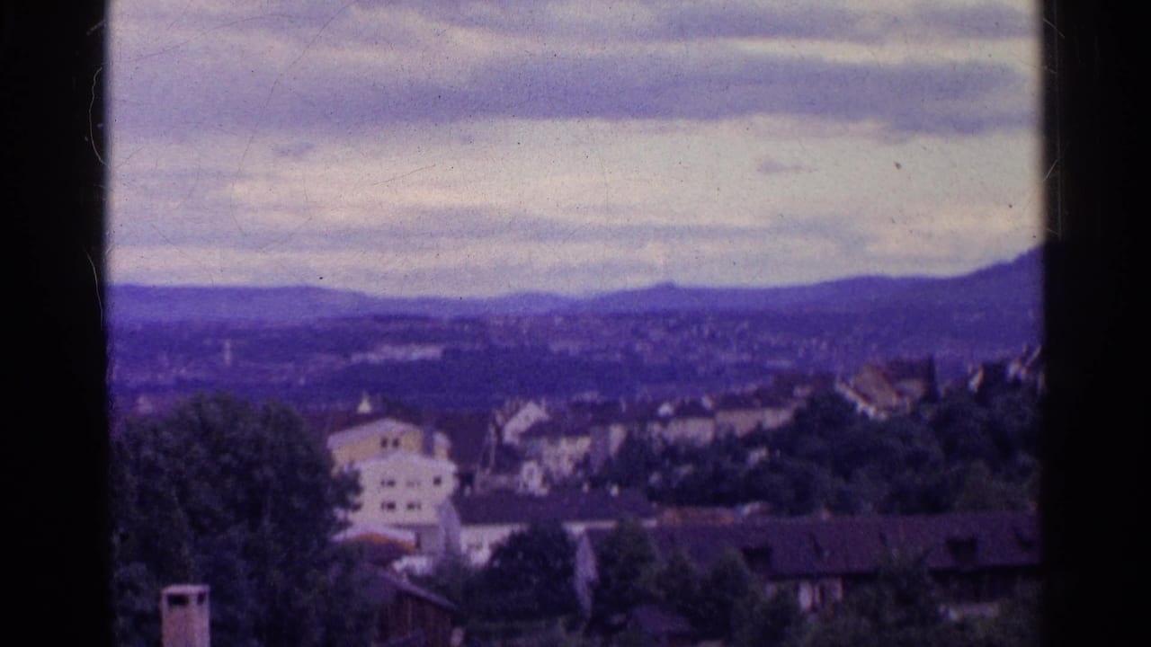 1961 Swiss home-movie frame with houses and trees in the foreground and a hazy mountain ridge beneath overcast clouds.
