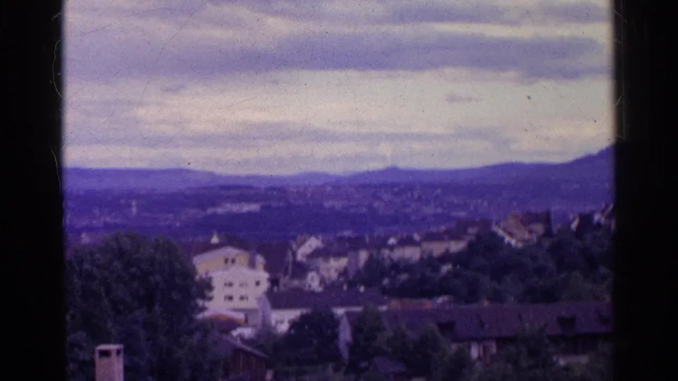 1961 Swiss home-movie frame with houses and trees in the foreground and a hazy mountain ridge beneath overcast clouds.