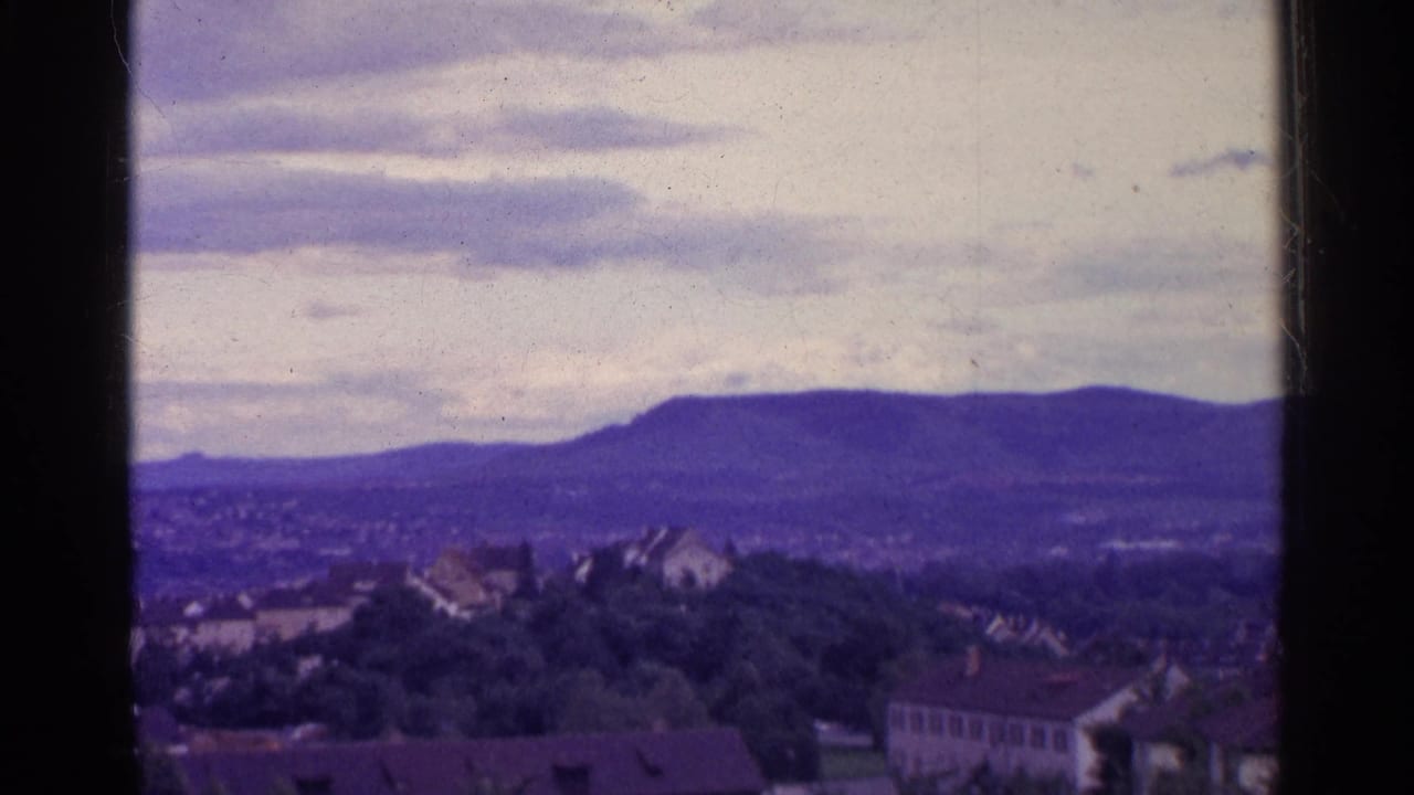 1961 Switzerland 8mm frame showing rooftops and wooded slopes leading toward distant hills under a bright overcast sky.