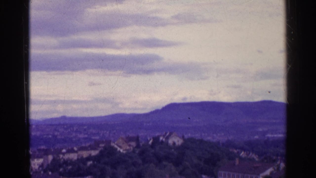 1961 Swiss 8mm frame of a town edge, trees, and a dark distant ridge beneath a pale cloudy sky.
