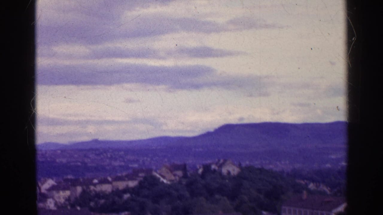 1961 8mm view of a Swiss valley town below distant mountains, framed by dark vertical edges and an overcast sky.