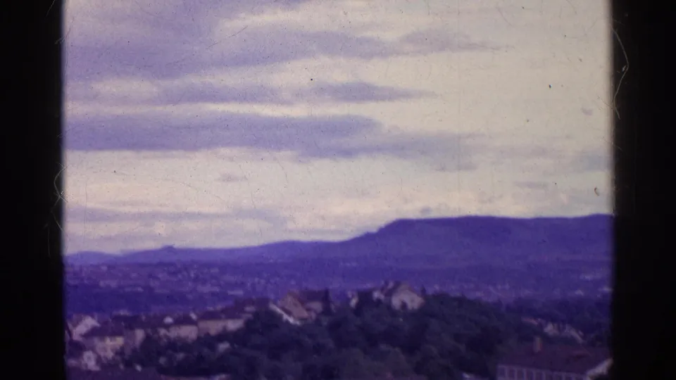 1961 8mm view of a Swiss valley town below distant mountains, framed by dark vertical edges and an overcast sky.