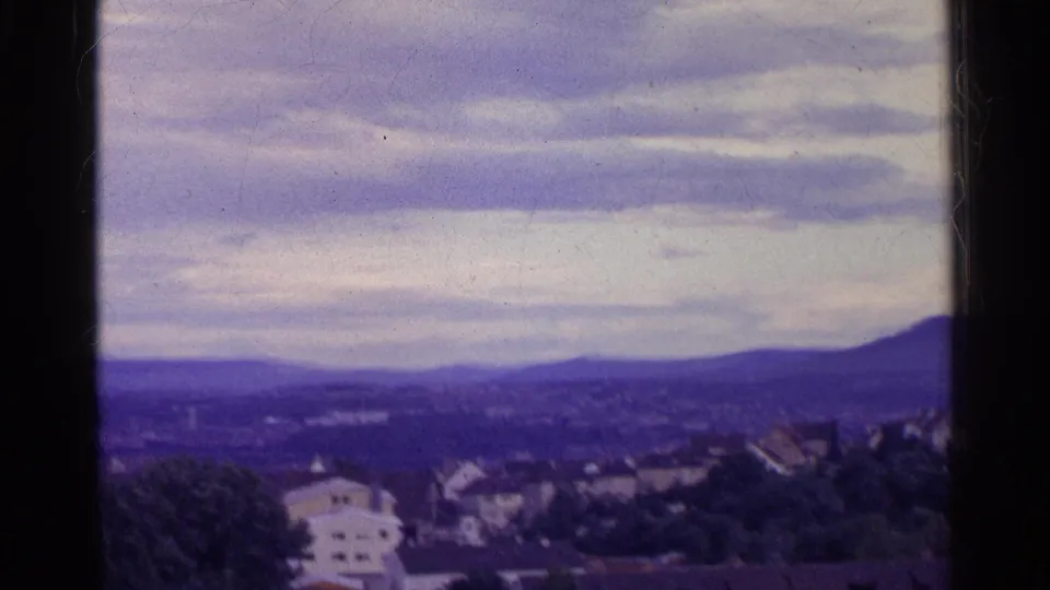 Swiss valley panorama from 1961 with rooftops, distant hills, layered clouds, and a dark vertical border crossing the view.
