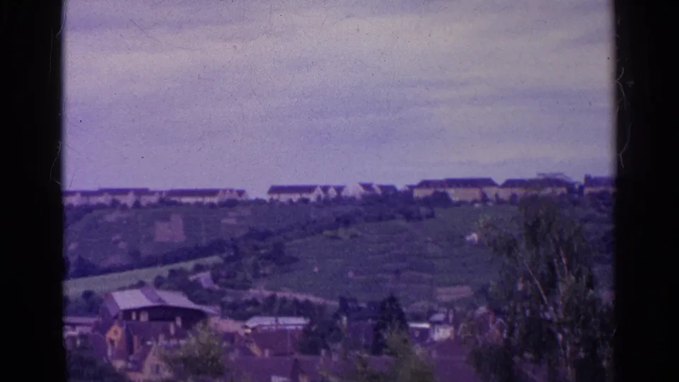 1961 8mm Swiss valley frame showing houses, green fields, trees, distant low buildings, and a pale cloudy sky.