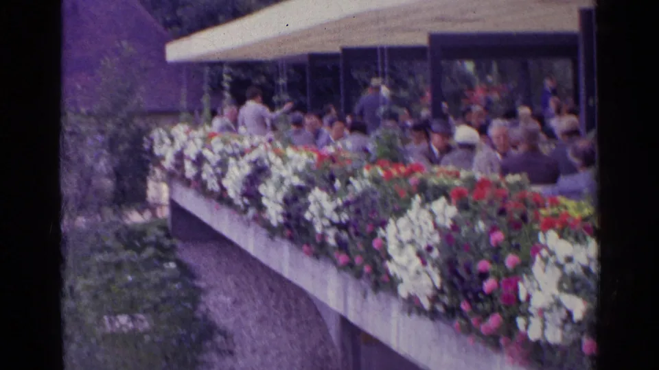 1961 Switzerland frame showing a flower-lined concrete planter, garden path and adults gathered beneath a covered pavilion.
