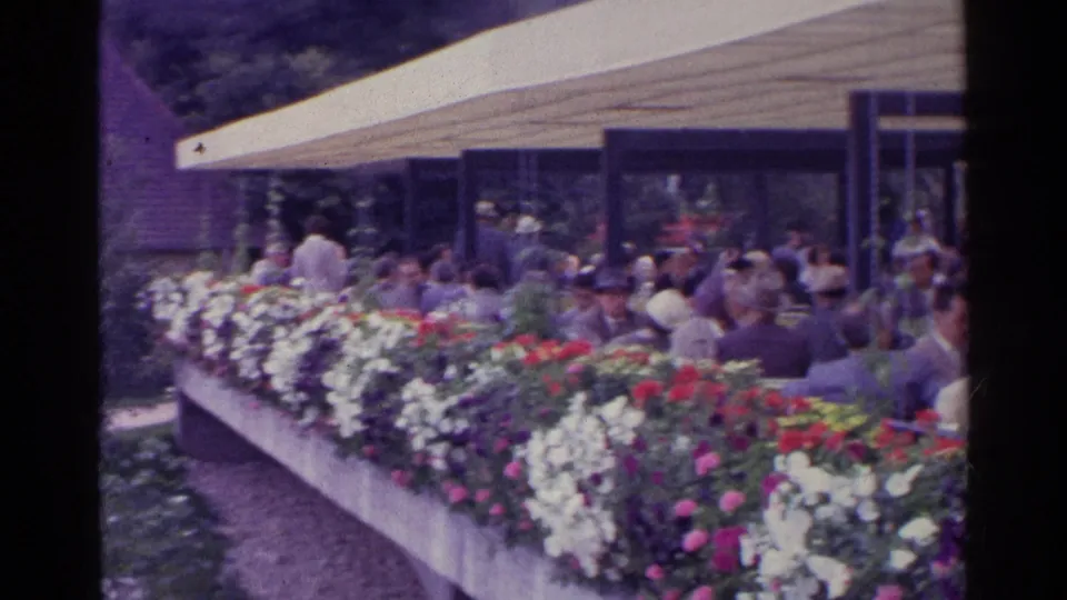 Home-movie frame from 1961 Switzerland showing a crowded covered terrace behind a long bed of colorful flowers.