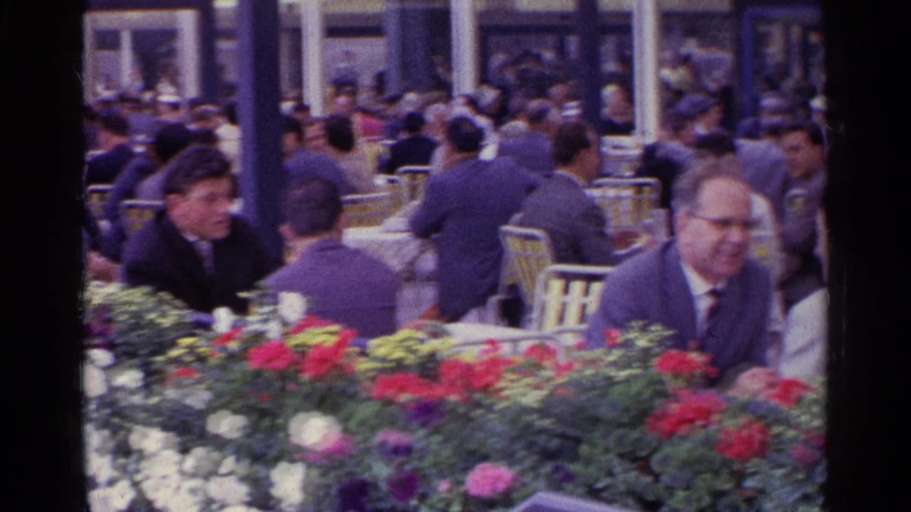 Swiss 1961 home-movie frame of cafe patrons in suits and hats seated beside a thick foreground flower bed.