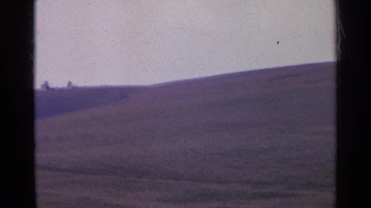 1961 Swiss home-movie frame of a sloping grass hill, with tiny distant figures or structures near the ridge.