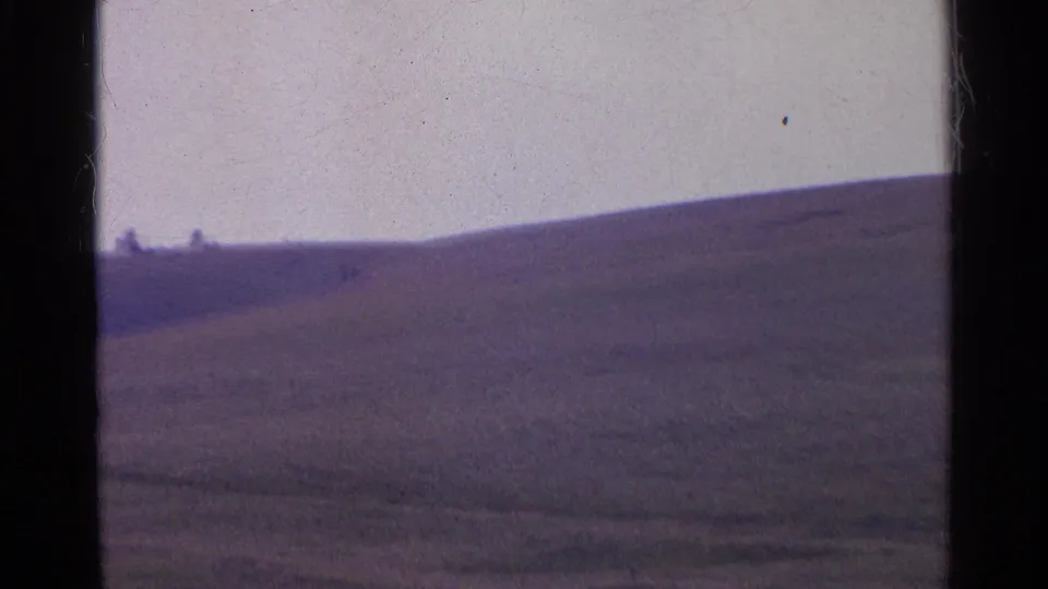 1961 Swiss home-movie frame of a sloping grass hill, with tiny distant figures or structures near the ridge.