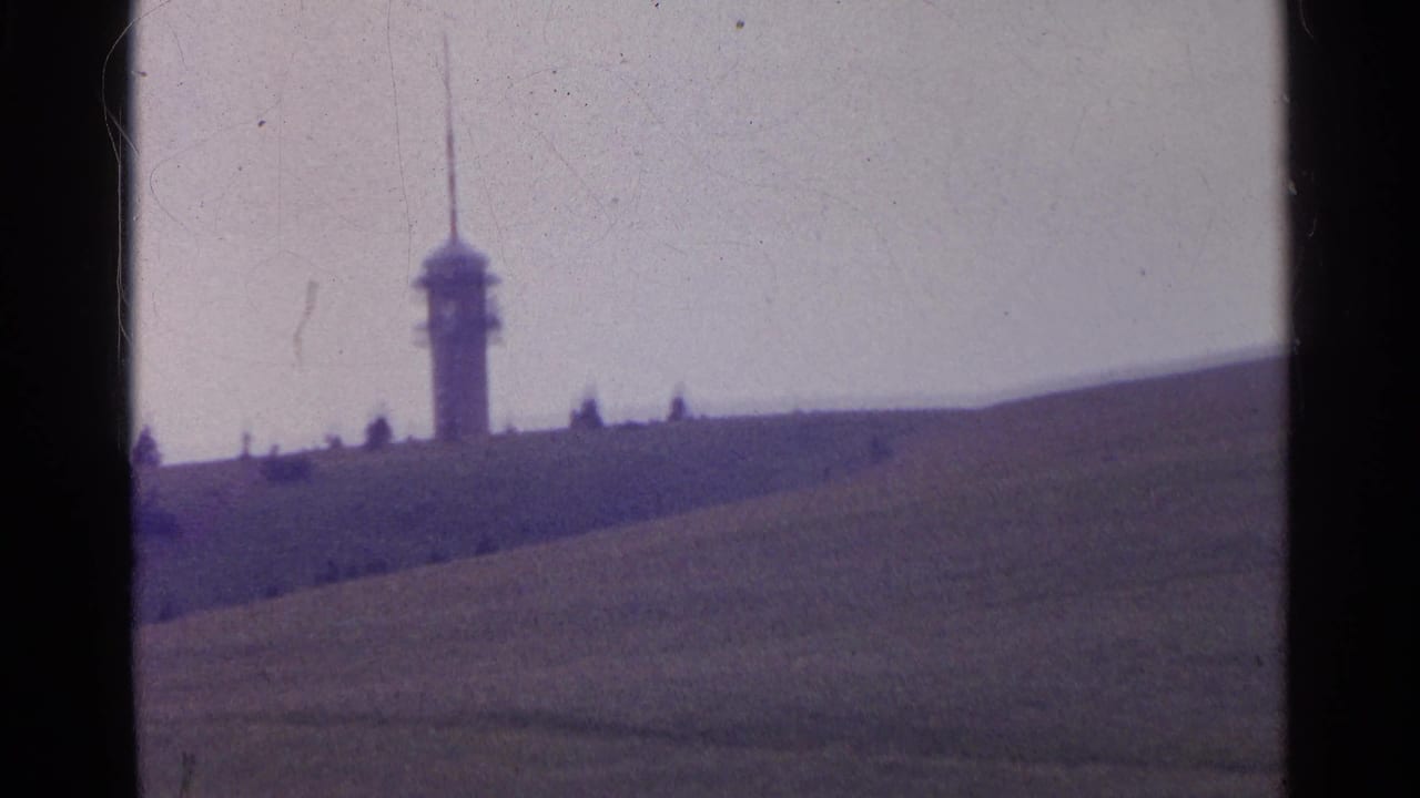 Distant ridge-top tower with a spire in 1961 Swiss 8mm footage, seen across rolling grassland with small visitors nearby.