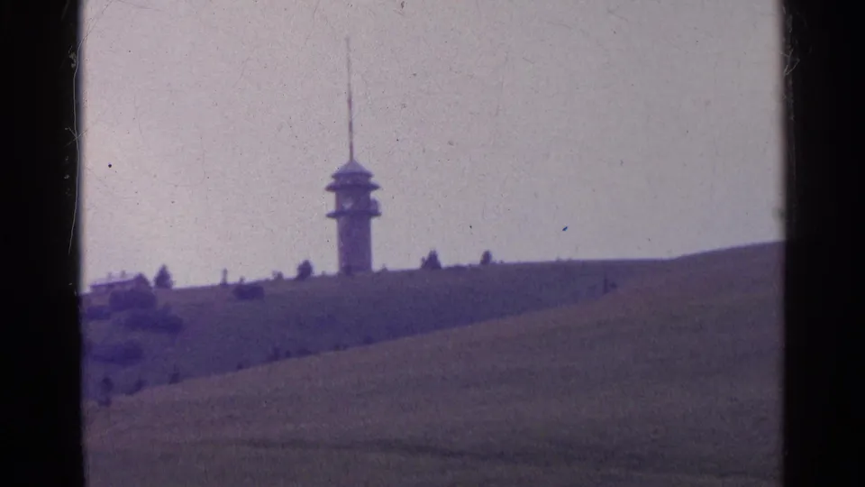 1961 Switzerland home-movie frame showing a slender distant tower on a hill with rolling green fields and sparse trees.