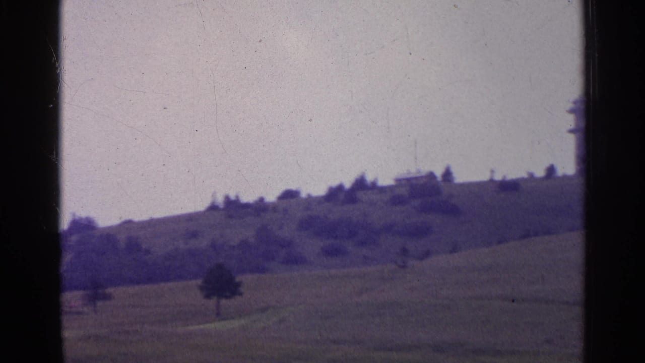 1961 Switzerland landscape frame with sloping pasture, scattered trees, and part of a distant tower near the frame edge.