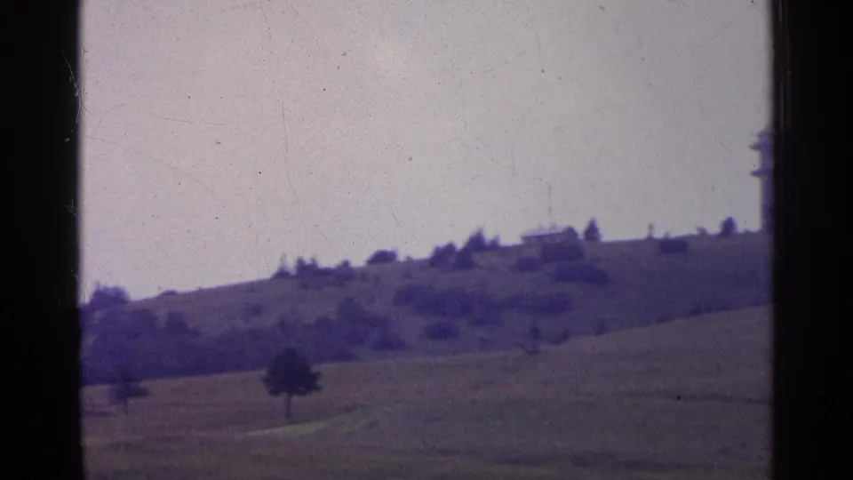 1961 Switzerland landscape frame with sloping pasture, scattered trees, and part of a distant tower near the frame edge.