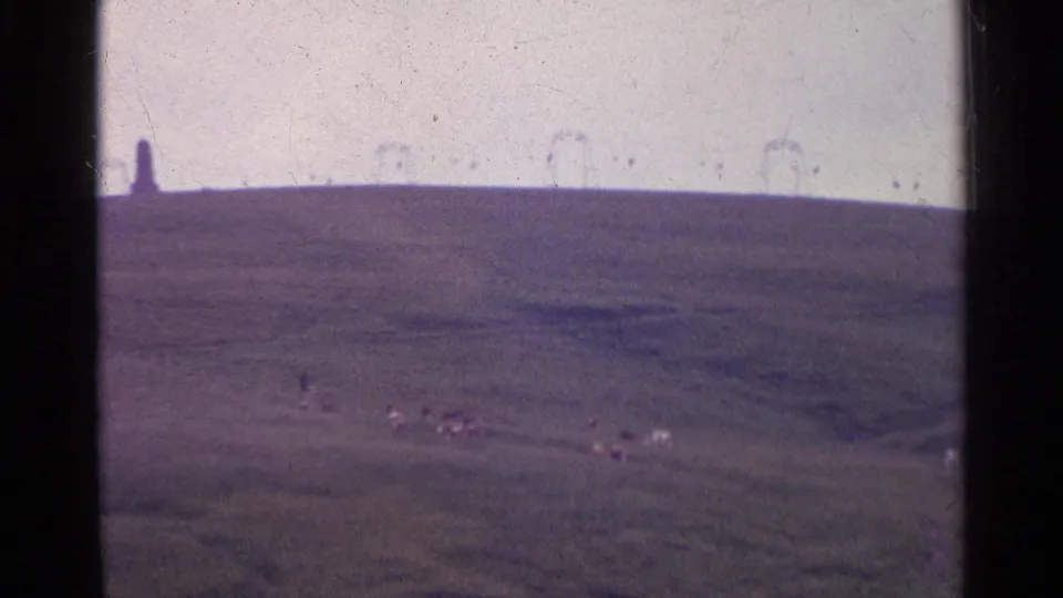 1961 Swiss countryside frame with grazing sheep on a sloping field and a distant monument-like shape along the ridge.
