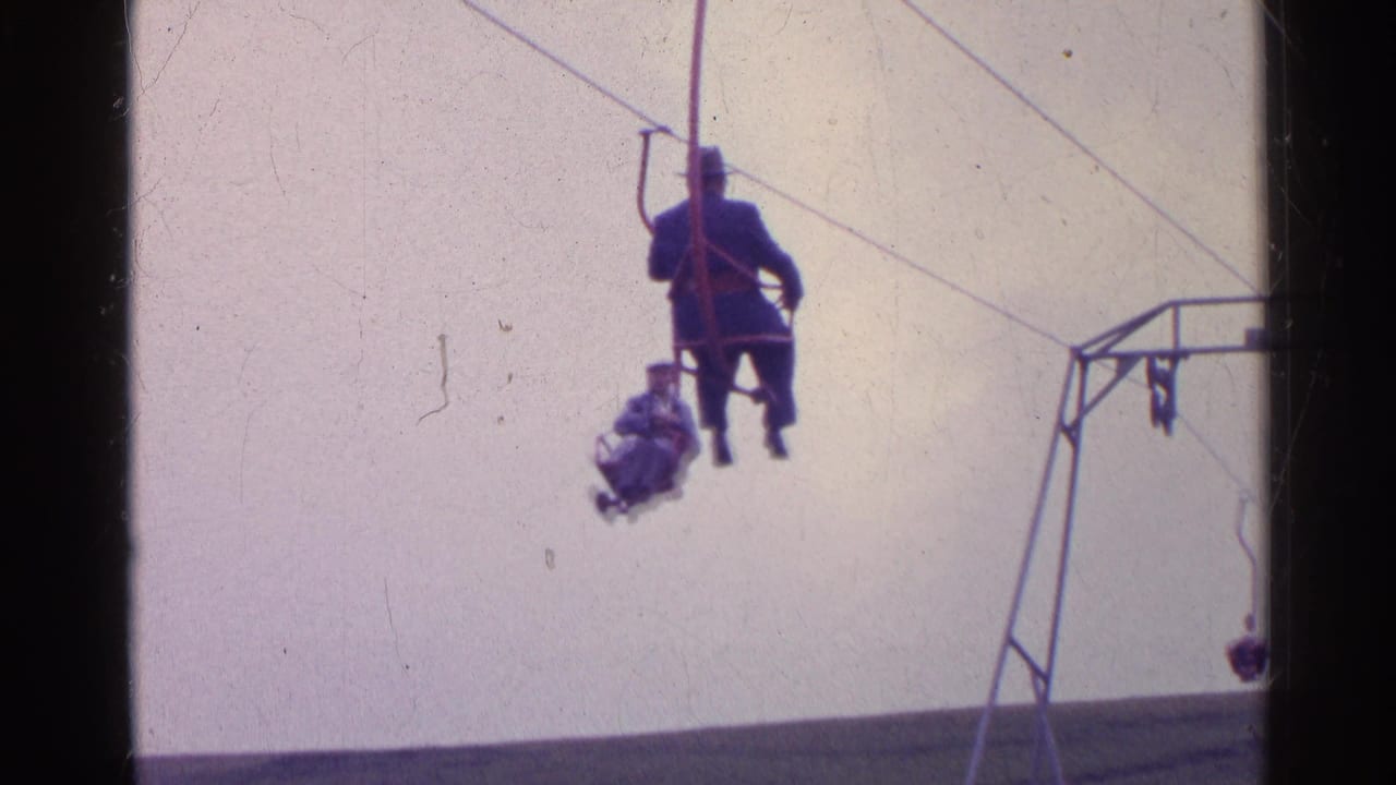 1961 Switzerland frame with distant riders on a small ski lift crossing a pale sky above a grassy alpine slope.