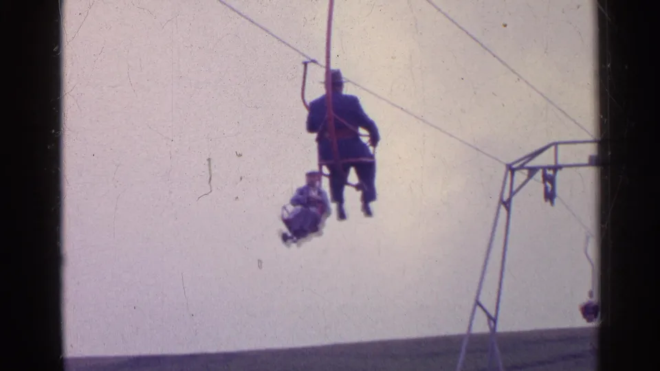 1961 Switzerland frame with distant riders on a small ski lift crossing a pale sky above a grassy alpine slope.