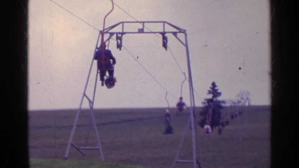 1961 Swiss home-movie frame showing a small chairlift with riders ascending over a grassy hill under a pale sky.