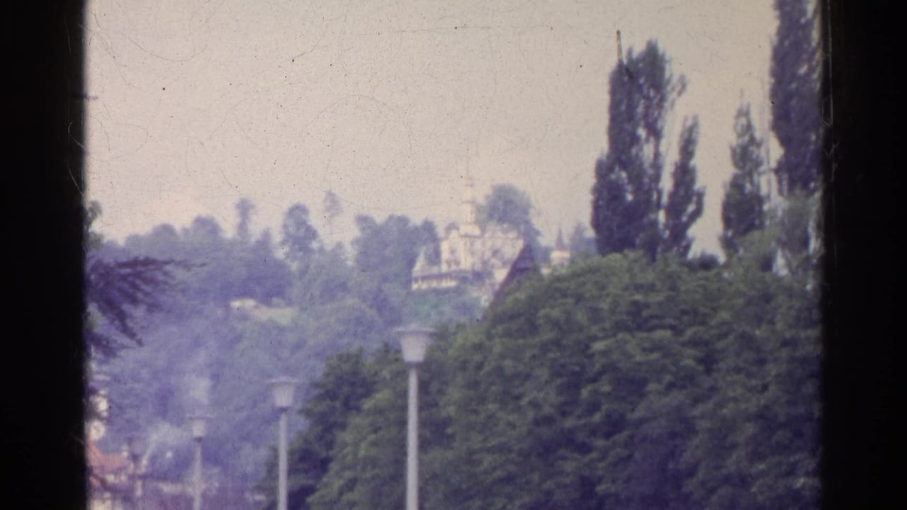 1961 Swiss travel footage looking across leafy treetops and street lamps toward distant hillside buildings.