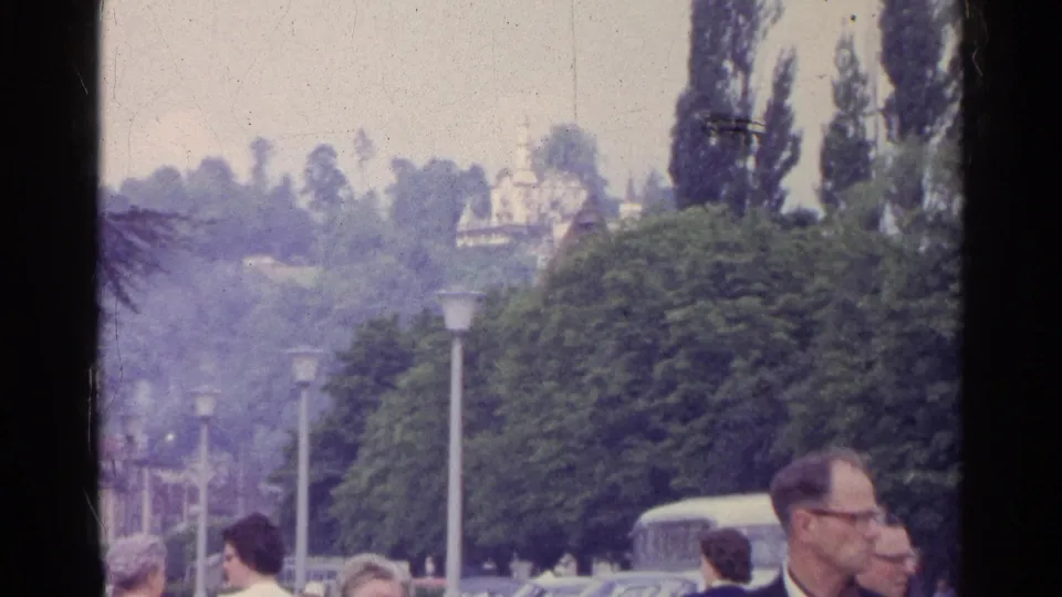 Swiss town street view with pedestrians near a bus, leafy trees, street lamps, and distant hillside architecture.