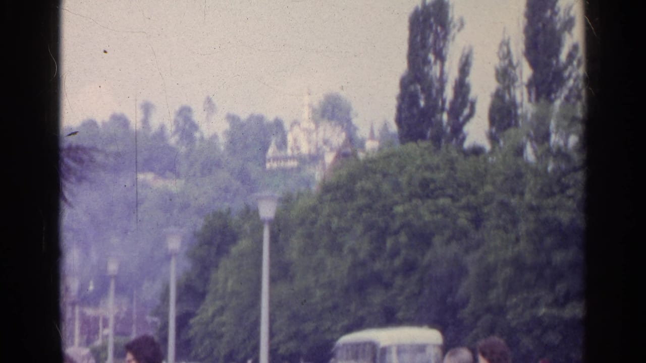 1961 Switzerland home-movie frame showing trees, street lamps, distant cars, and a pale classical-style building near a fountain.