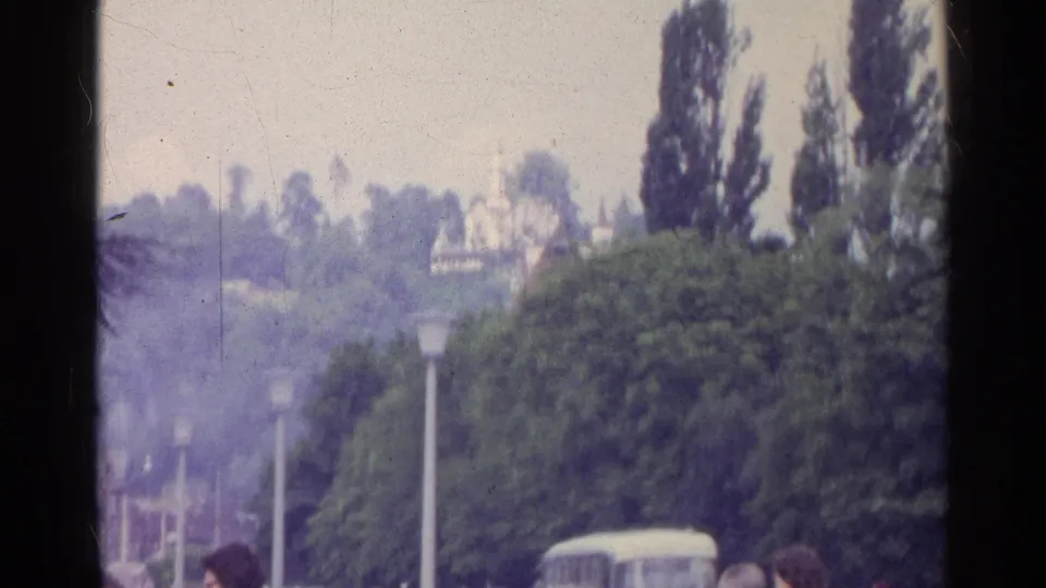 1961 Switzerland home-movie frame showing trees, street lamps, distant cars, and a pale classical-style building near a fountain.