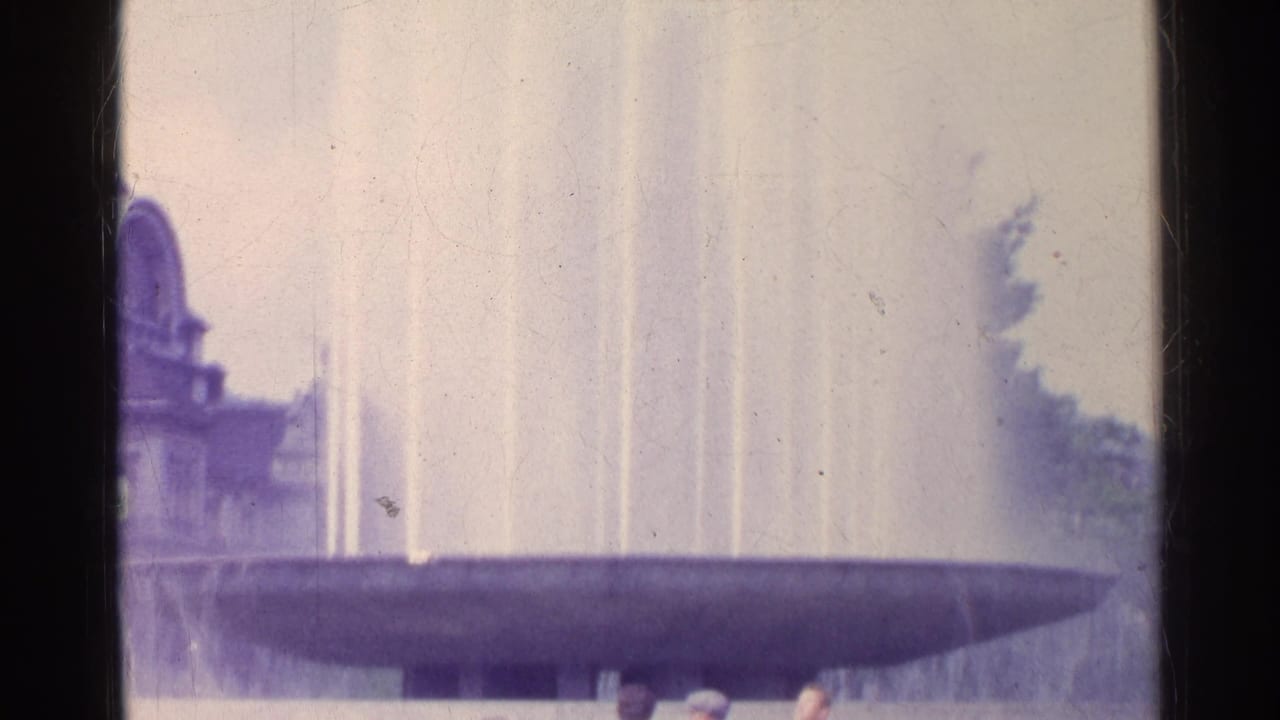 1961 Switzerland home-movie frame showing a large public fountain with vertical jets and a partial classical-style building.