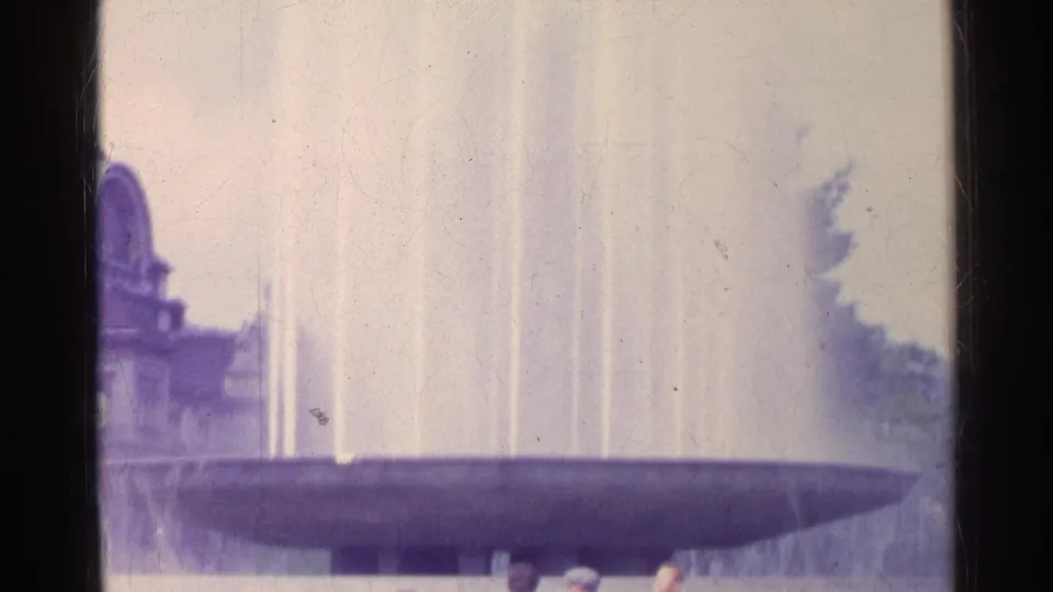 1961 Switzerland home-movie frame showing a large public fountain with vertical jets and a partial classical-style building.