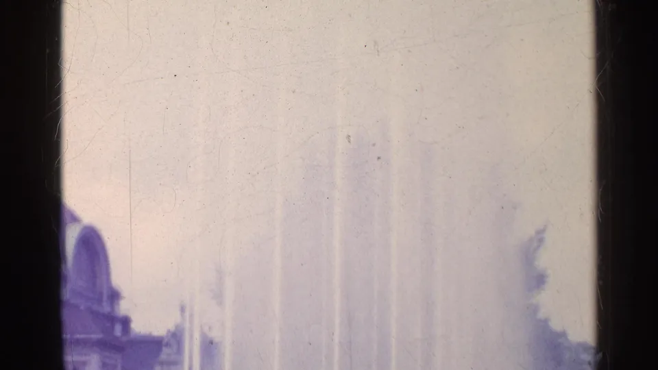 1961 Swiss 8mm frame showing pale fountain jets rising beside a partly visible arched public building.