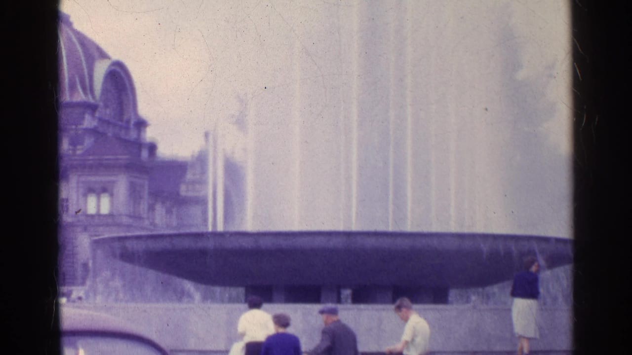 1961 Swiss 8mm frame showing a broad public fountain basin, tall jets, arched building, and visitors in period dress.