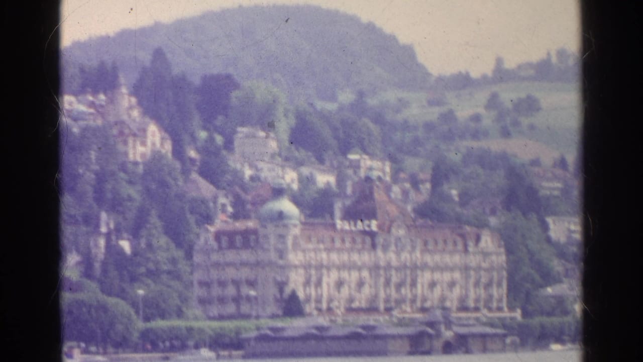 Swiss lakefront in 1961 with a prominent PALACE hotel, hillside residences, trees, and soft faded 8mm color.