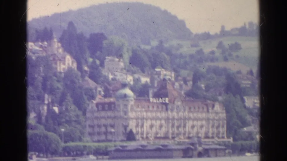 Swiss lakefront in 1961 with a prominent PALACE hotel, hillside residences, trees, and soft faded 8mm color.