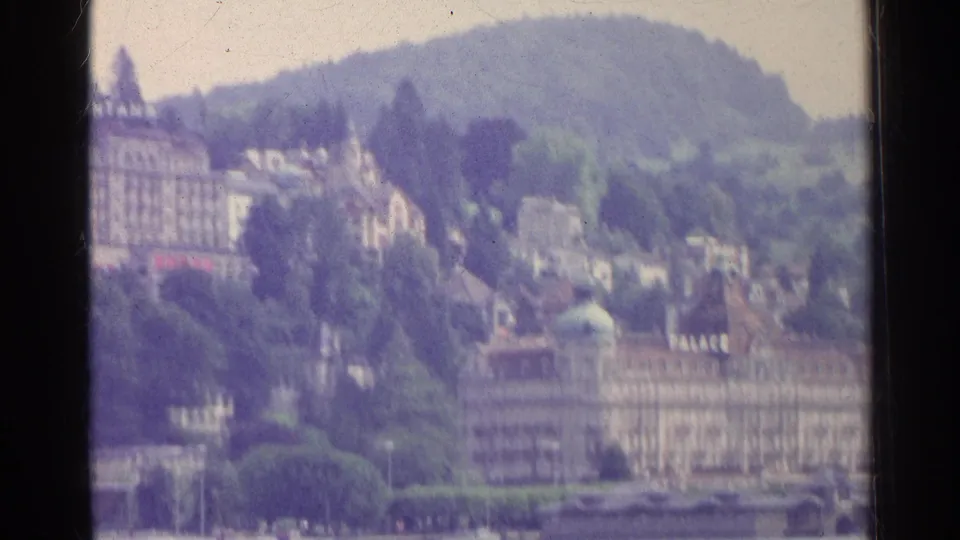 1961 Swiss home-movie frame with lakeside hotel architecture, hillside homes, trees, and the lake foreground.