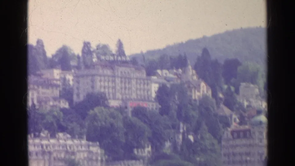 1961 Swiss lakeside view showing a hotel with a MONTANA sign, wooded slopes, hillside buildings, and soft 8mm grain.
