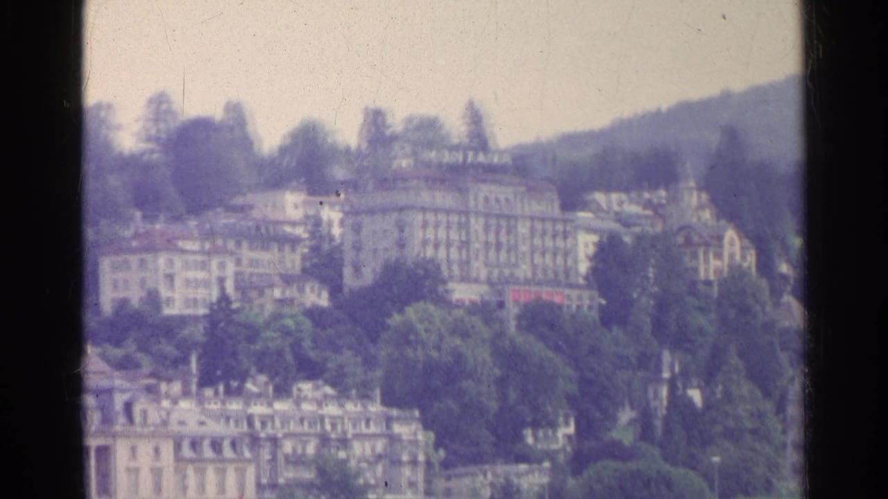1961 Swiss lakeside view with hillside hotels, trees, and a waterfront casino building seen in soft 8mm focus.