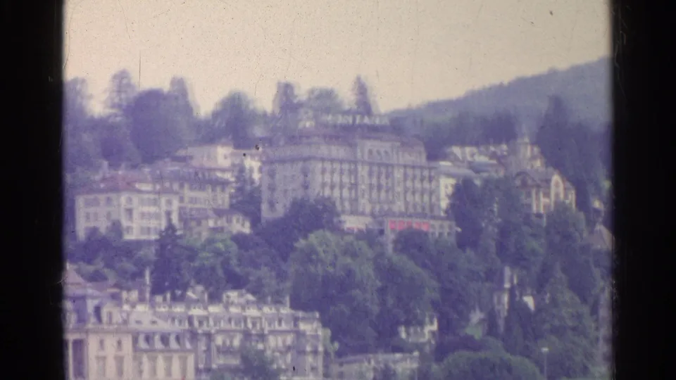 1961 Swiss lakeside view with hillside hotels, trees, and a waterfront casino building seen in soft 8mm focus.