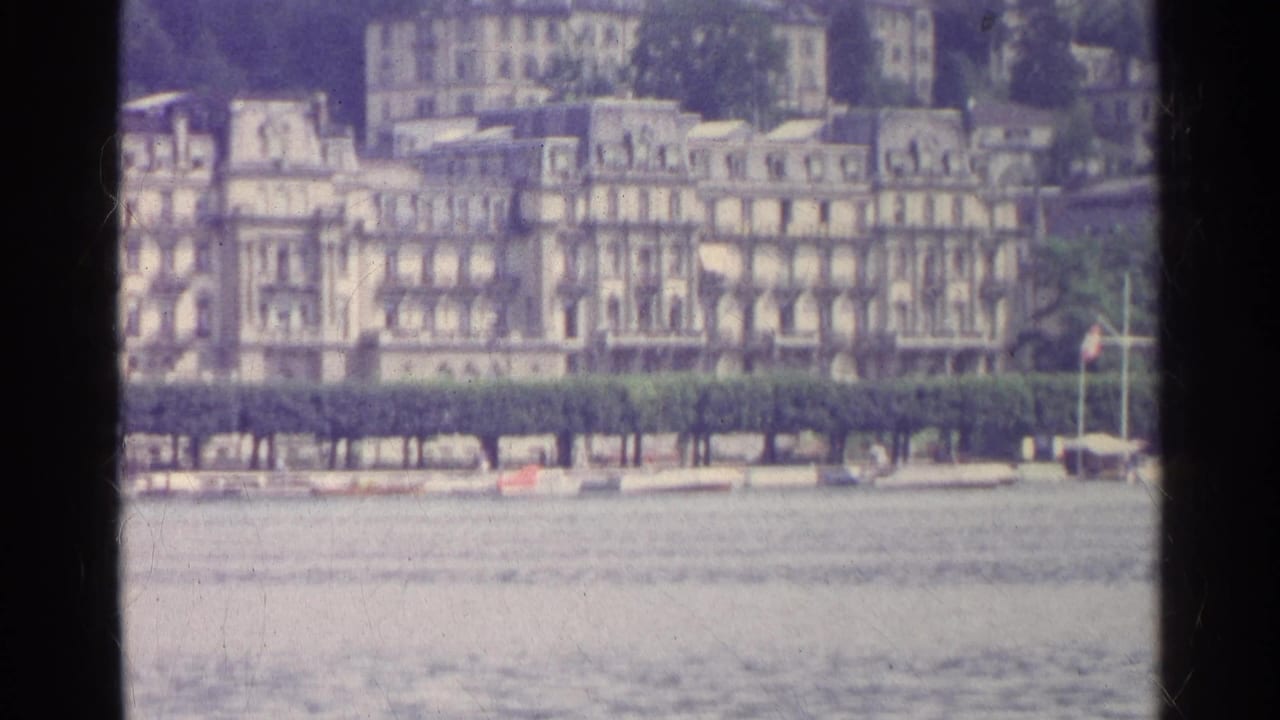 1961 Swiss lakeside scene with a large resort building, arched windows, flags, trees, hills, and water in the foreground.