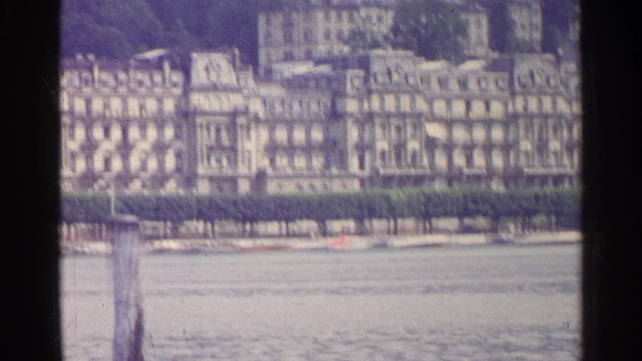 1961 Swiss 8mm frame showing distant ornate lakefront buildings behind a hedge, with water and a wooden post in the foreground.