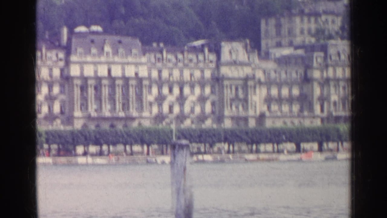 Row of large waterfront buildings behind a hedge across calm water.