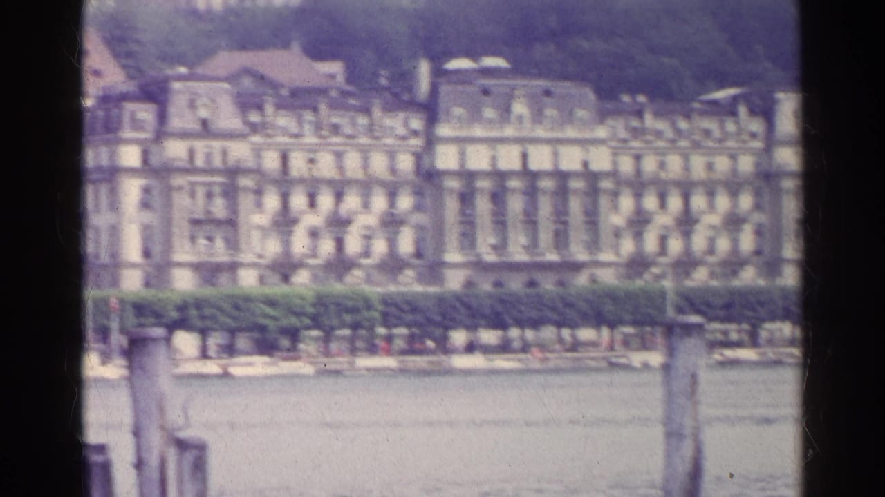 1961 Swiss home-movie frame of pale classical buildings across a lake, with a hedge and foreground wooden post.