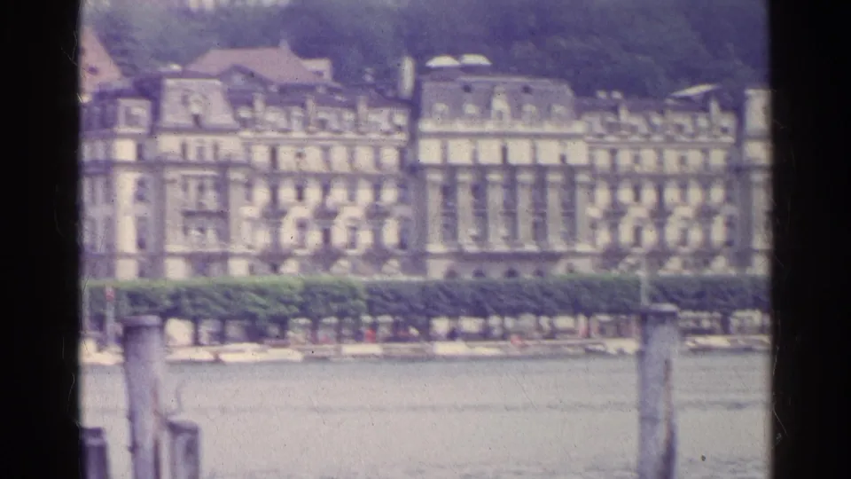 1961 Swiss home-movie frame of pale classical buildings across a lake, with a hedge and foreground wooden post.