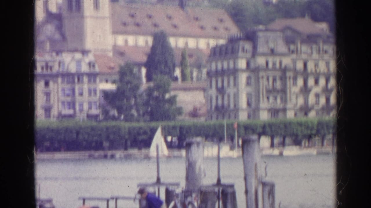 1961 Swiss lakeside view with a church tower, waterfront buildings, hedges, posts, and small boats or docks in the foreground.