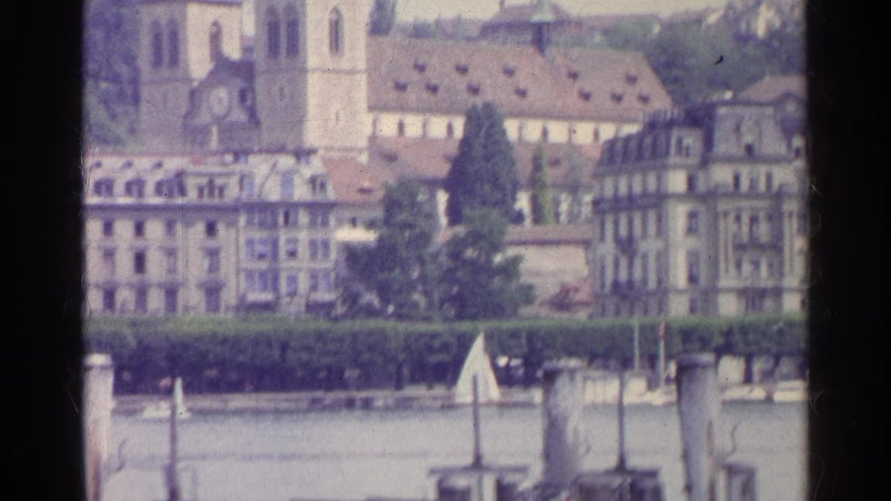 1961 8mm view of Swiss lakeside buildings with a twin-spired church, tiled rooftops, trees, and waterfront structures.